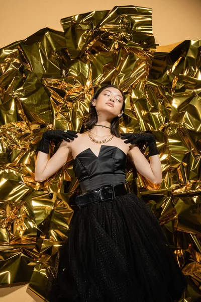 Elegant asian young woman with short hair and closed eyes posing in black strapless dress with tulle skirt and gloves while standing next to shiny background, model, wrinkled golden foil — Stock Photo