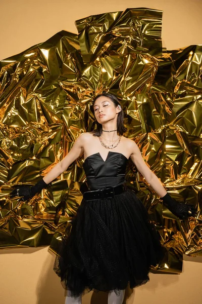 Enchanting asian young woman with short hair posing in black strapless dress with tulle skirt and gloves while standing next to shiny yellow background, model, wrinkled golden foil, looking away — Stock Photo