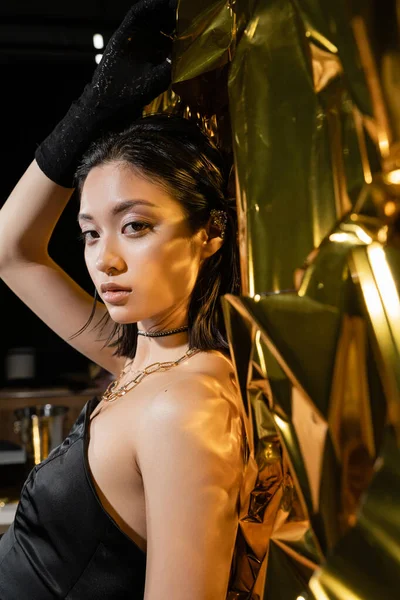 Asian young woman with wet short hair posing in strapless dress with black glove and ear cuff while standing next to shiny background, model, looking at camera, wrinkled golden foil — Stock Photo