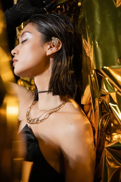 Asiatische junge Frau mit nasser Frisur und kurzen Haaren posiert im trägerlosen Kleid mit schwarzem Handschuh und Ohrenmanschette neben glänzendem Hintergrund, Modell, geschlossene Augen, faltige Goldfolie — Stockfoto