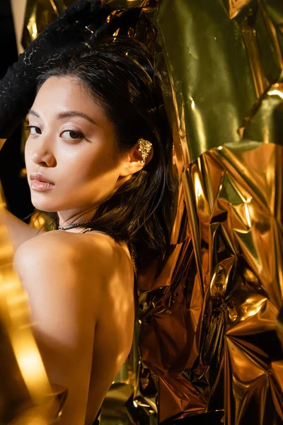 Elegant asian young woman with wet hairstyle and short hair, shimmer eyeshadow posing in black glove while standing next to shiny background, model, looking at camera, wrinkled golden foil — Stock Photo