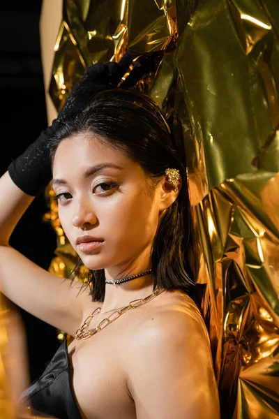 Elegante asiático jovem mulher com molhado penteado e curto cabelo posando em strapless vestido com preto luva enquanto em pé ao lado de ouro fundo, modelo, olhando para câmera, enrugado amarelo folha — Fotografia de Stock