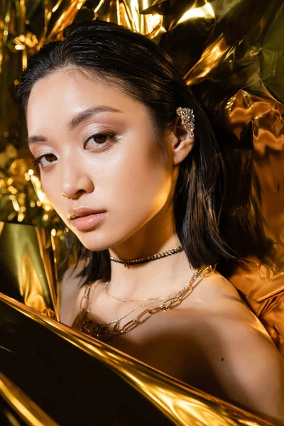 Retrato de hermosa mujer asiática joven con el pelo corto mojado posando junto a fondo brillante, modelo, mirando a la cámara, lámina dorada arrugada, maquillaje natural, belleza asiática - foto de stock