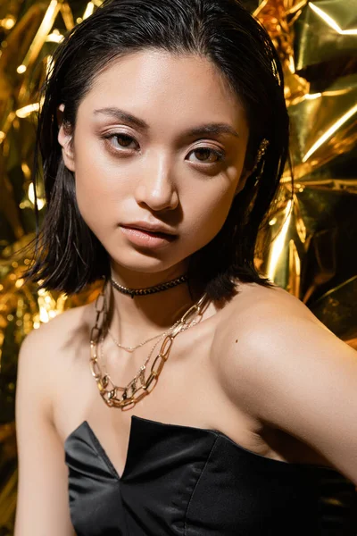 Beautiful asian young woman with wet hairstyle and short hair posing in strapless dress with black glove while standing next to shiny golden background, model, looking at camera, wrinkled golden foil — Stock Photo