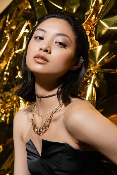 Beautiful asian young woman with short hair posing in strapless dress with black glove while standing next to shiny yellow background, model, looking at camera, wrinkled golden foil, wet hairstyle — Stock Photo