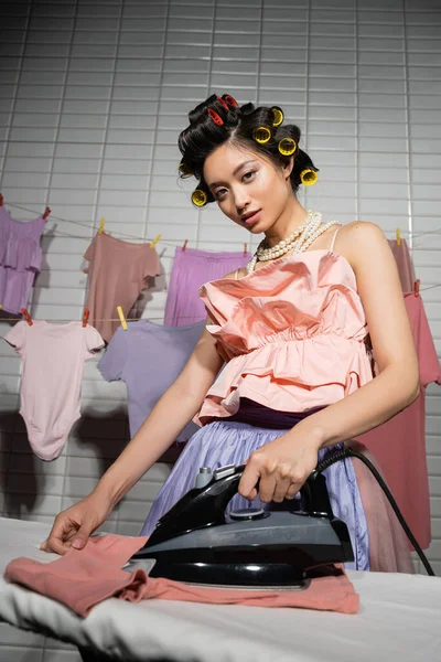 Baixo ângulo vista de jovem asiático dona de casa com cabelo encrespadores engomar enquanto em pé em rosa babados topo e pérola colar perto de limpar roupas penduradas no fundo borrado, mulher, lavanderia, limpeza — Fotografia de Stock