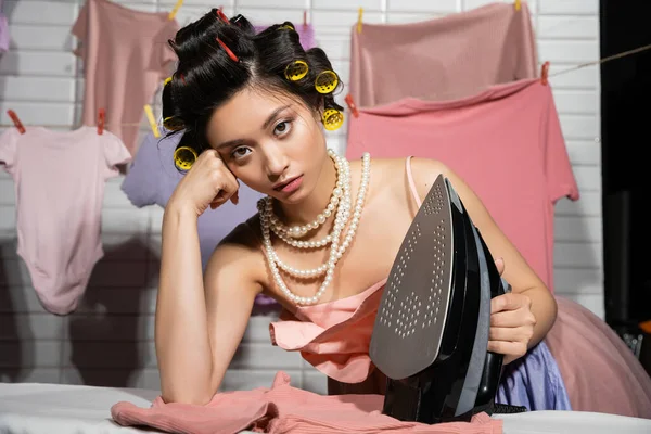 Müde asiatische Hausfrau mit Lockenwicklern in rosa Rüschenoberteil und Perlenkette, die in die Kamera schaut, während sie Eisen in der Nähe sauberer Kleidung hält, die auf verschwommenem Hintergrund hängt, Hausarbeit, junge Frau, Wäsche — Stockfoto