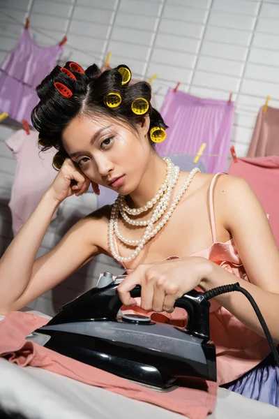 Tired asian housewife with hair curlers in pink ruffled top and pearl necklace ironing and looking at camera near wet clothes hanging on blurred background, housework, young woman, laundry — Stock Photo