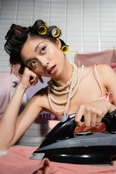 Мечтательная азиатская домохозяйка с волосами и жемчужное ожерелье гладильная и глядя в сторону около чистой и мокрой одежды висит на размытом фоне, домашняя работа, молодая женщина, прачечная — стоковое фото
