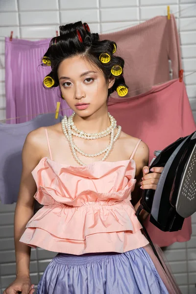 Asian young woman with hair curlers holding iron while posing in pink ruffled top and pearl necklace near clean and wet laundry hanging on blurred background, housework, housewife — Stock Photo
