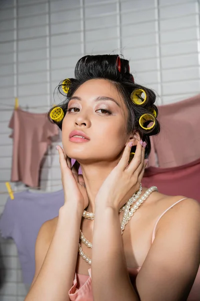 Молодая азиатская женщина касаясь волос с бигуди волос и позируя в жемчужном ожерелье около чистой и влажной прачечной висит на размытом фоне, домашняя работа, домохозяйка, глядя в камеру — стоковое фото