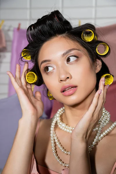 Mujer asiática joven pensativa tocando el pelo con rulos de pelo y posando en collar de perlas cerca de ropa limpia y húmeda colgando sobre fondo borroso, tareas domésticas, ama de casa, mirando hacia otro lado - foto de stock