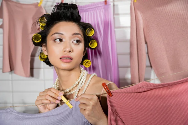 Задумчивая молодая азиатская женщина с бигуди волос и жемчужное ожерелье с булавкой одежды и висит чистое и влажное белье с размытым фоном на дому, домашняя работа, домохозяйка, домашние обязанности — стоковое фото