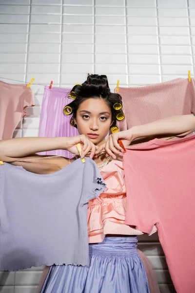 Young asian woman with hair curlers posing in ruffled top and pearl necklace near clean and wet clothes hanging in laundry room, housework, housewife, looking at camera, domestic responsibilities — Stock Photo