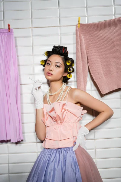 Asiatische junge Frau mit Lockenwicklern in rosa Rüschenoberteil, Perlenkette und Handschuhen, während sie Zigarette neben nasser Wäsche hält, die neben weißen Fliesen hängt, Hausfrau, in die Kamera schauend, rauchend — Stockfoto