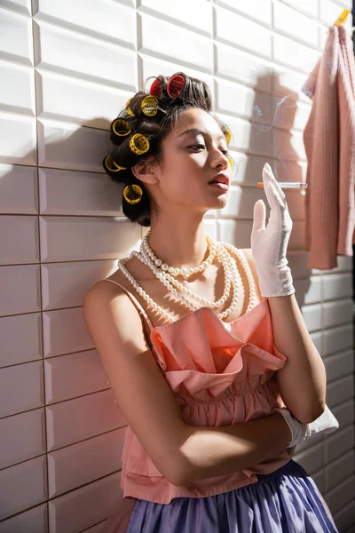 Mujer joven asiática con rizadores de pelo de pie en rosa con volantes superior, collar de perlas y guantes mientras sostiene el cigarrillo cerca de ropa mojada colgando cerca de azulejos blancos, ama de casa, mirando hacia otro lado, fumando - foto de stock