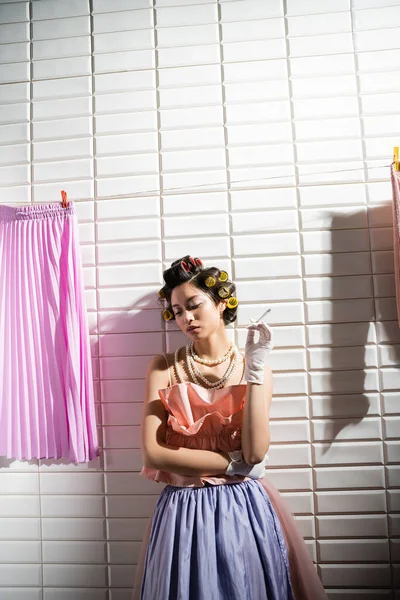 Asian young woman with hair curlers standing in pink ruffled top, pearl necklace and gloves while holding cigarette near wet laundry hanging near white tiles, housewife, smoking, bad habit — Stock Photo