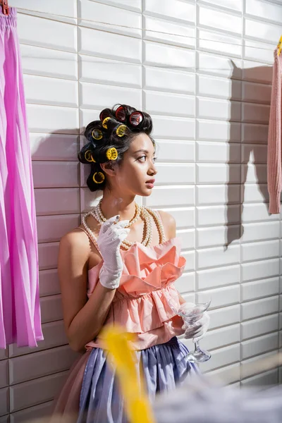 Asian young woman with hair curlers standing in pink ruffled top, pearl necklace and gloves smoking and holding glass near wet laundry hanging near white tiles, iron, cigarette, housewife — Stock Photo