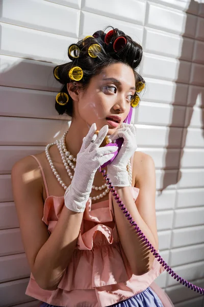 Brunette and asian young woman with hair curlers standing in pink ruffled top, pearl necklace and white gloves, smoking cigarette and talking on retro phone near white tiles, housewife, vintage — Stock Photo