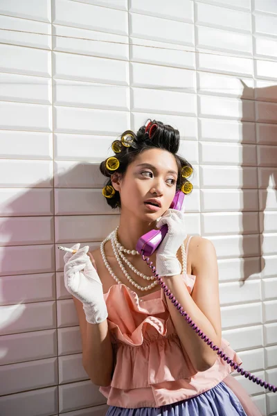 Fashionable and asian young woman with hair curlers standing in pink ruffled top, pearl necklace and white gloves smoking and talking on retro phone near white tiles, housewife, holding cigarette — Stock Photo