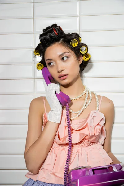 Brunette and asian young woman with hair curlers standing in pink ruffled top, pearl necklace and white gloves and talking on purple retro phone near white tiles, housewife, looking away — Stock Photo
