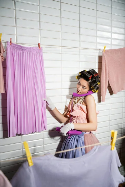 Asian young woman with hair curlers standing in pink ruffled top, pearl necklace and white gloves, talking on purple retro phone and looking at wet skirt near clean laundry handing near white tiles — Stock Photo