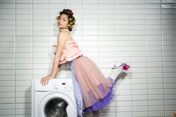 Mujer joven asiática con rizadores de pelo de pie en rosa con volantes superior, collar de perlas, falda de tul y zapatos de tacón alto con pluma cerca de la lavadora moderna cerca de azulejos blancos en el cuarto de lavado - foto de stock