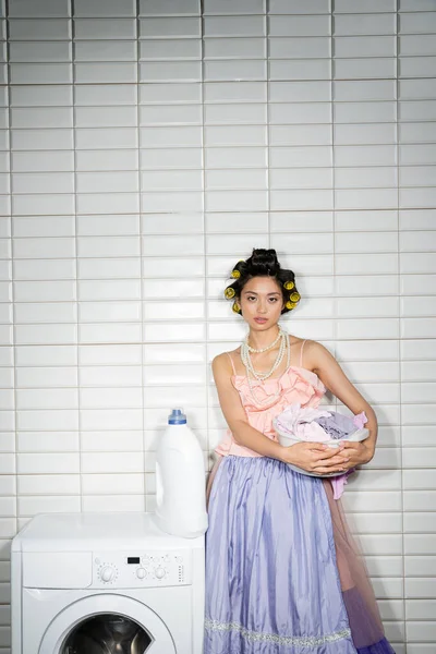 Asian young woman with hair curlers standing in pink ruffled top, pearl necklace and tulle skirt and holding washing bowl with dirty clothes near modern washing machine and detergent in laundry room — Stock Photo