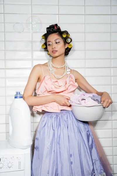 Asian young woman with hair curlers standing in pink ruffled top, pearl necklace, tulle skirt and holding washing bowl with dirty clothes near washing machine and detergent in laundry room, soap bubbles — Stock Photo