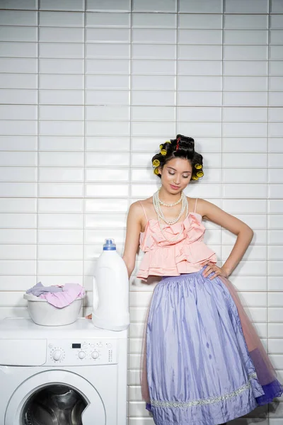 Asiática jovem com encrespadores de cabelo de pé com a mão no quadril em babados top, colar de pérolas e saia de tule perto de bacia de lavagem com roupas sujas na máquina de lavar roupa moderna com detergente na lavanderia — Fotografia de Stock