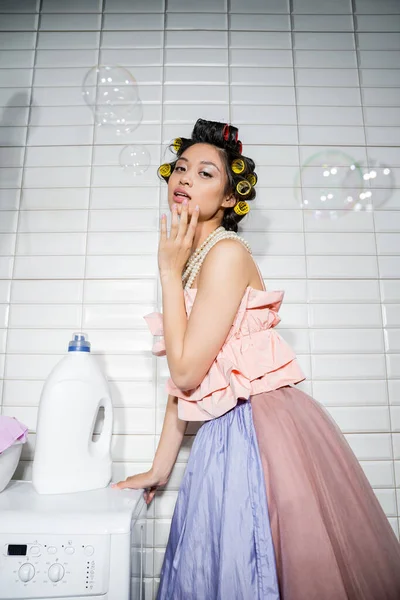 Young asian woman with hair curlers standing in ruffled top, pearl necklace and tulle skirt near modern washing machine with detergent bottle in laundry room, housewife, soap bubbles — Stock Photo