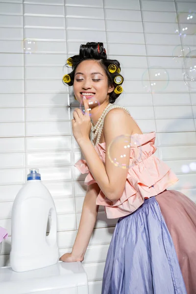 Happy and young asian woman with hair curlers standing in ruffled top, pearl necklace and tulle skirt near modern washing machine with detergent bottle in laundry room, housewife, soap bubbles — Stock Photo