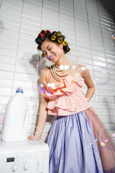 Happy and young asian woman with hair curlers standing in ruffled top, pearl necklace and tulle skirt near detergent bottle on modern washing machine in laundry room, housewife, soap bubbles — Stock Photo