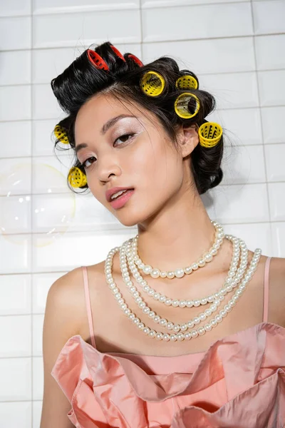 Portrait de brune et jeune femme asiatique avec des boucles de cheveux debout dans collier de perles près de bulle de savon floue dans la buanderie avec des carreaux blancs, femme au foyer, beauté naturelle — Photo de stock