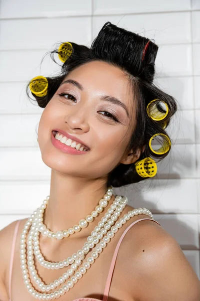 Ritratto di donna asiatica allegra e giovane con bigodini in piedi in collana di perle e sorridente in lavanderia con piastrelle bianche, casalinga, bellezza naturale — Foto stock