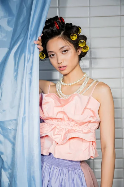 Brunette and asian young woman with hair curlers standing in pink ruffled top with pearl necklace near blue bathroom curtain and looking at camera near white tiles at home — Stock Photo