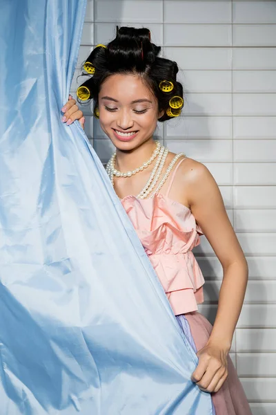 Heureux et asiatique jeune femme avec des bigoudis de cheveux debout en haut à volants rose avec collier de perles près bleu rideau de salle de bains et regarder vers le bas près de tuiles blanches à la maison — Photo de stock