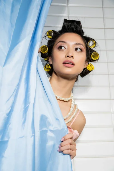 Neugierige und asiatische junge Frau mit Lockenwicklern, die in Perlenkette hinter blauem Badezimmervorhang steht und in der Nähe weißer Fliesen zu Hause wegschaut, Hausfrau, häusliche Szene — Stockfoto