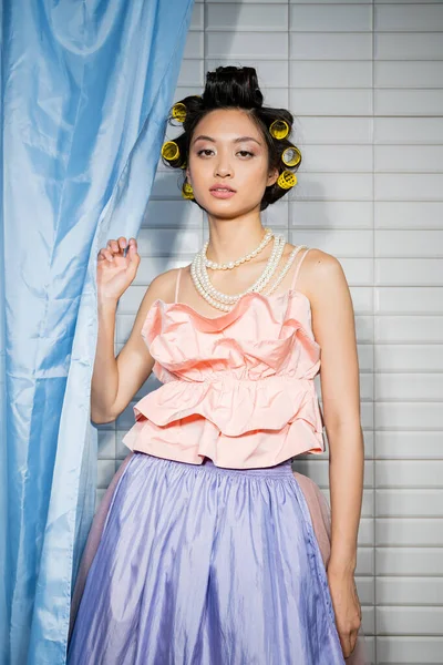 Mujer joven de moda y asiática con rizadores de pelo de pie en rosa con volantes superior con collar de perlas cerca de la cortina de baño azul y mirando a la cámara cerca de azulejos blancos en casa - foto de stock