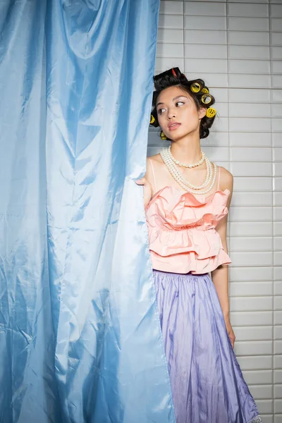 Brunette and asian young woman with hair curlers standing in pink ruffled top with pearl necklace and skirt near blue bathroom curtain and looking away near white tiles at home — Stock Photo