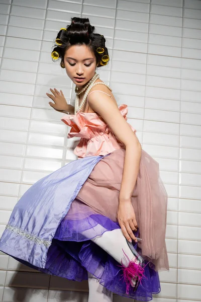 Low angle view of asian young woman with hair curlers standing in pink ruffled top, pearl necklace, skirt and feather heels while posing near white tiles in bathroom — Stock Photo