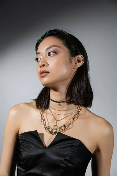 Retrato de morena e asiática jovem mulher com cabelo curto posando em vestido sem alças preto olhando afastado no fundo cinza, colares, maquiagem natural, penteado molhado — Fotografia de Stock