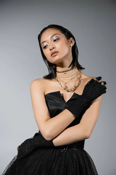Portrait of brunette and asian young woman with short hair posing in black strapless dress and gloves with golden rings, looking away on grey background, necklaces, natural makeup — Stock Photo