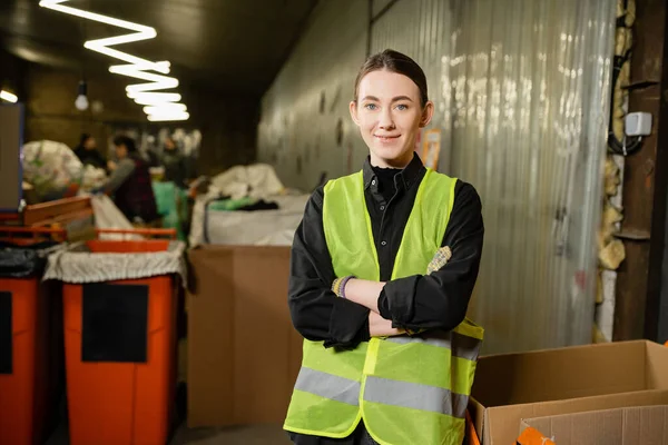 Young and smiling worker of waste disposal station in high visibility vest looking at camera and crossing arms while standing near blurred bins at background, garbage recycling concept — Stock Photo