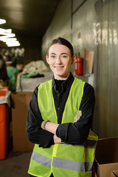 Positive and young worker in gloves and high visibility jacket crossing arms and looking at camera while standing in blurred waste disposal station at background, garbage sorting and recycling concept — Stock Photo