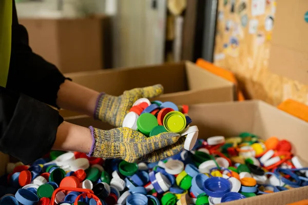 Ausgeschnittene Ansicht einer Sortiererin in Schutzhandschuhen, die bunte Plastikverschlüsse in der Nähe von Kartons hält, während sie in der Müllentsorgungsstation arbeitet, Müllsortier- und Recyclingkonzept — Stockfoto