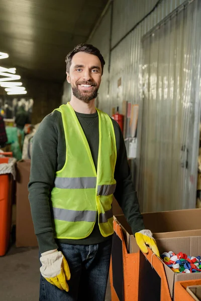 Cheerful sorter in high visibility vest and protective gloves looking at camera while standing near plastic caps in carton boxes in waste disposal station, garbage sorting and recycling concept — Stock Photo