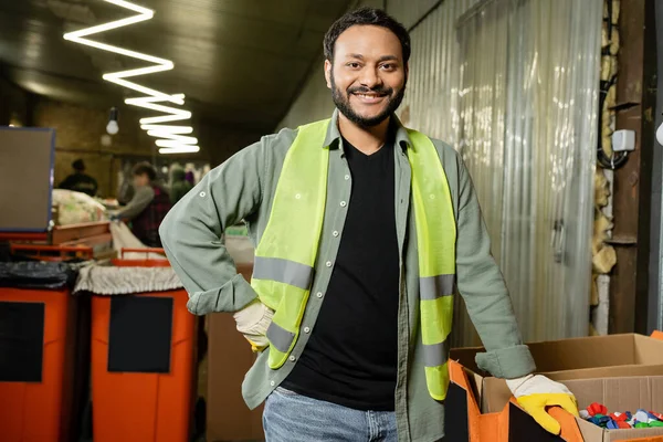 Smiling indian worker in high visibility vest and protective gloves looking at camera near plastic caps in carton boxes while working in waste disposal station, garbage sorting and recycling concept — Stock Photo