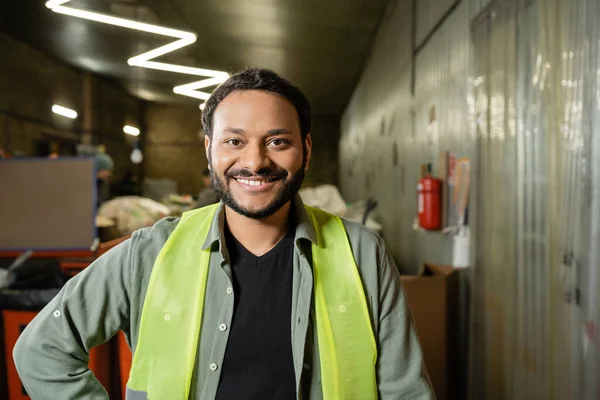 Portrait of cheerful indian sorter in high visibility vest looking at camera while standing and working in blurred waste disposal station, garbage sorting and recycling concept — Stock Photo