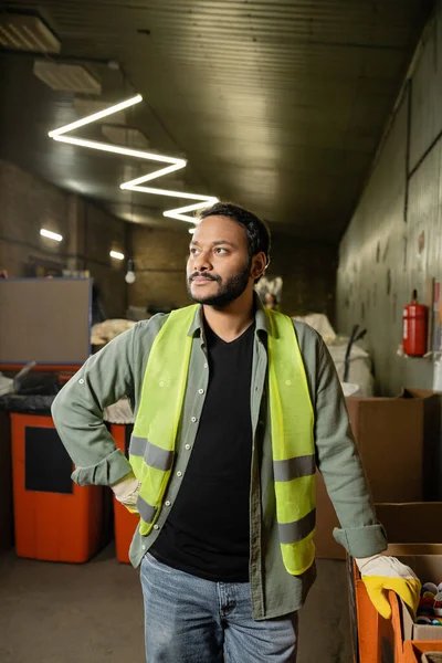 Indian man in high visibility vest and gloves looking away while standing near plastic caps in carton box and working in garbage sorting center, garbage sorting and recycling concept — Stock Photo